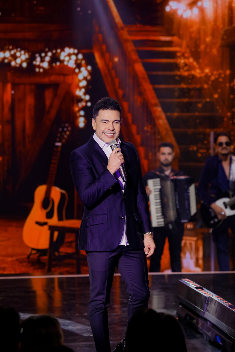 Zezé Di Camargo em pé e sorridente no palco do especial de Natal do SBT cantando 